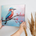 Framed canvas print of a colourful Kingfisher bird on a branch with pink cherry blossoms against a soft blue sky