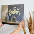 Framed canvas print of white flowers in glass vase with vintage bottle on a neutral background