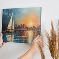 Framed canvas print of an impressionist painting with a sailboat against a sunset cityscape reflecting on the water