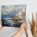 Framed canvas print of a realistic painting depicting a cliffside Mediterranean village with vivid blue sea and sky