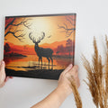 Printed canvas art of a black deer silhouette against an orange sunset with trees and mountains